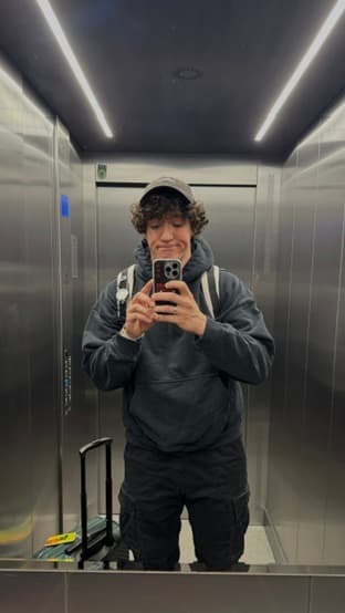 Kamil taking a mirror selfie in a steel elevator, suitcase at his feet